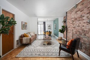 A modern living room with a brick wall, a wooden floor, a brown sofa, a coffee table, and a dining area in the background.