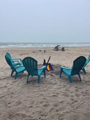Une plage avec trois chaises Adirondack turquoise disposées en demi-cercle autour d’un feu de camp.
