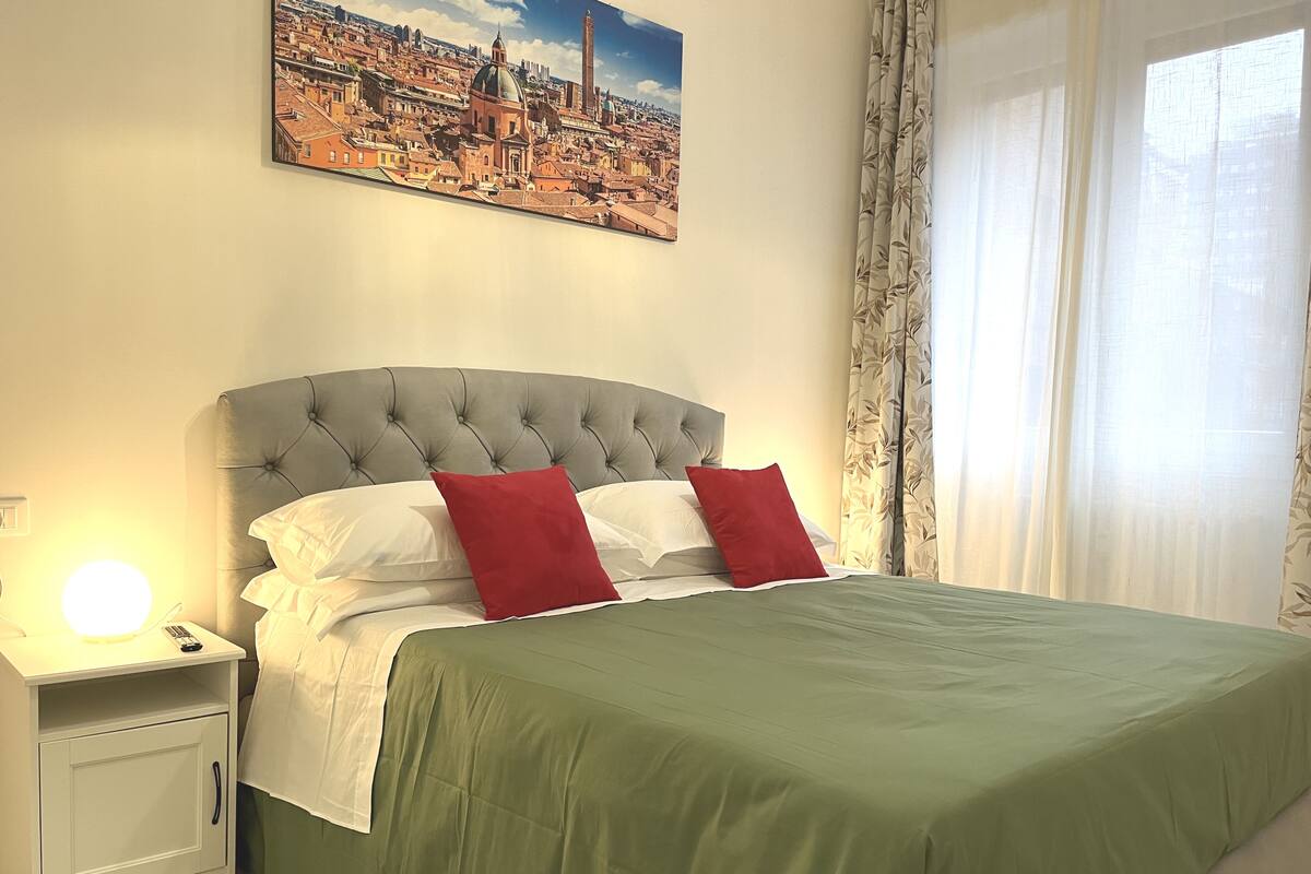 A neatly made bed with a green bedspread and two red pillows, a white bedside table with a lamp, and a cityscape painting on the wall.