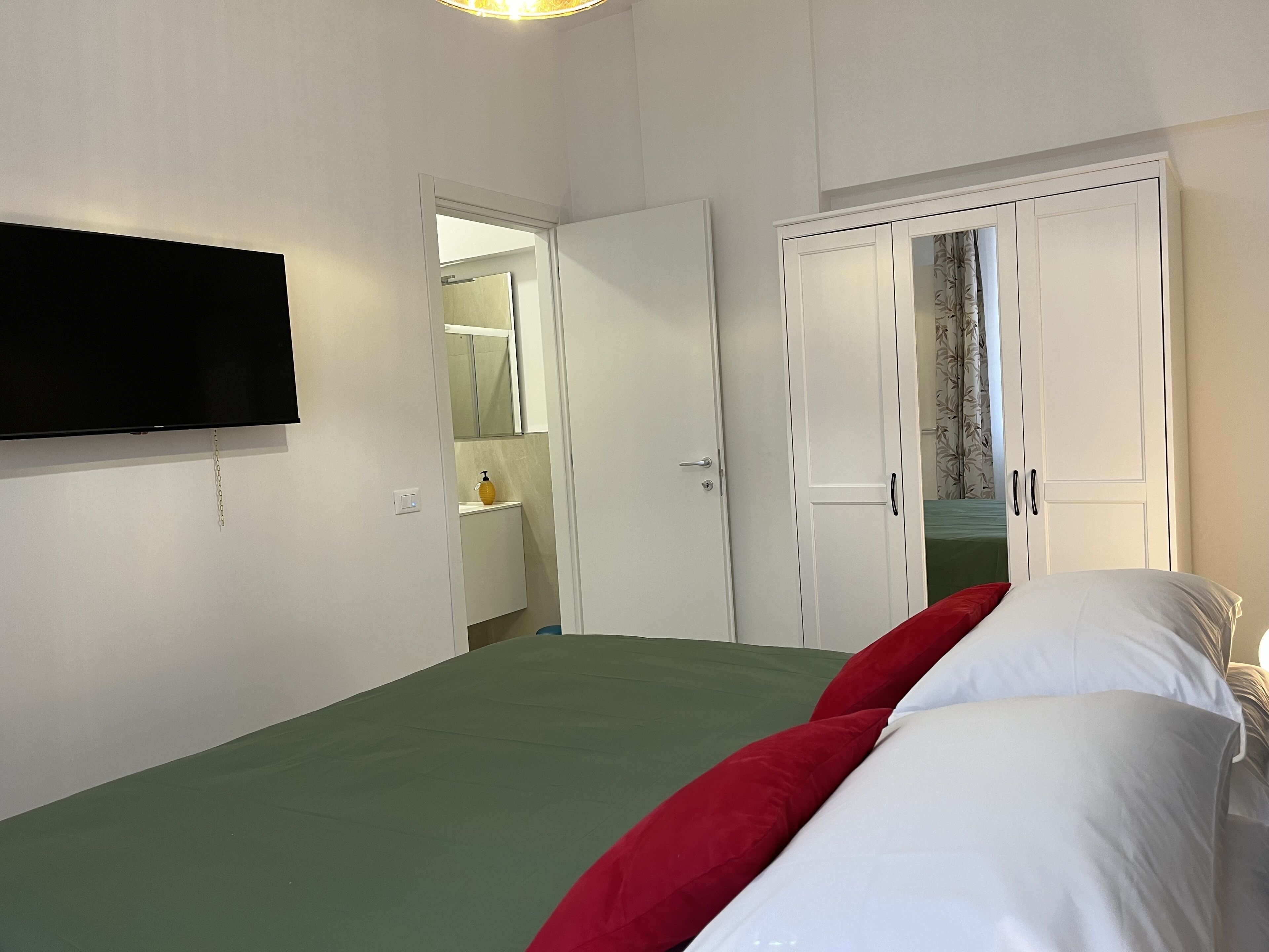 Une chambre à coucher avec un lit, un téléviseur fixé au mur, une armoire et une salle de bain visible à travers une porte ouverte.