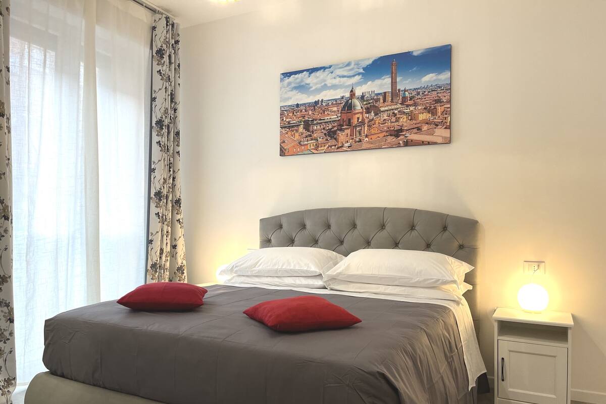 A bedroom with a bed, a nightstand, a wall-mounted lamp, and a cityscape painting.