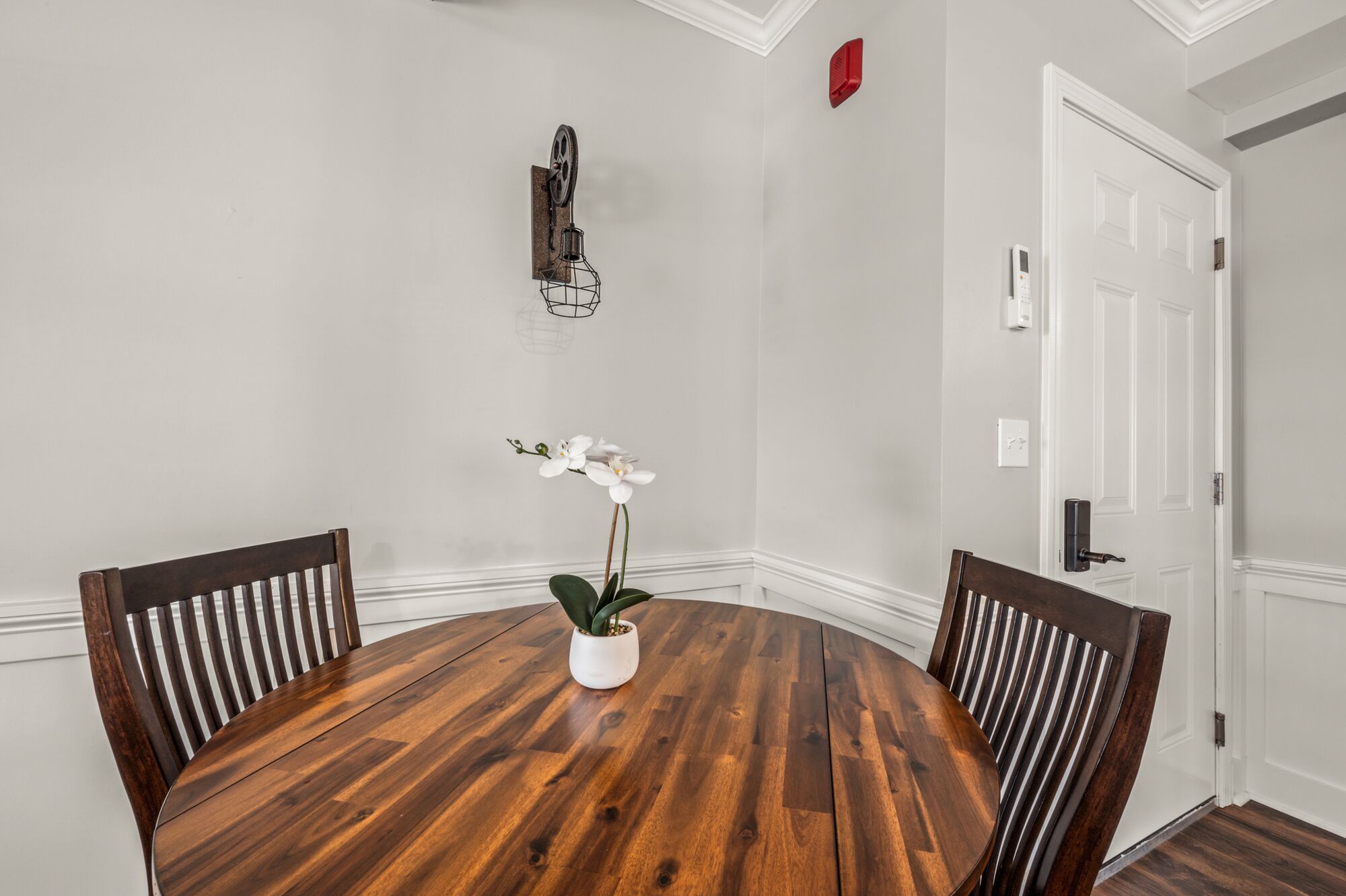 A round wooden dining table with four chairs, a white orchid in a pot, and a wall-mounted decorative piece.