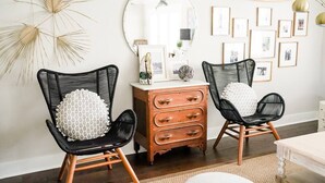 Una habitación con dos sillas de mimbre negras con cojines blancos, un aparador de madera, un espejo y una pared decorada con cuadros enmarcados.