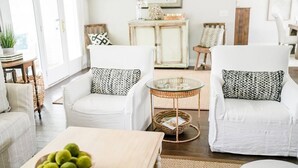 Una sala de estar acogedora con dos sillones blancos, una mesa redonda de vidrio y un cuenco de manzanas verdes sobre una mesa de madera.