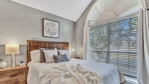 A bedroom with a large bed, a wooden headboard, a bedside table with a lamp, a framed picture on the wall, and a window with blinds. - Splash & Play Hideaway: Pool House + Game Room/Gym (Lewisville)