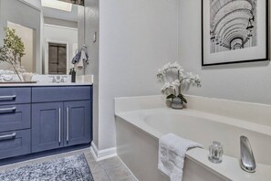 A modern bathroom with a large mirror, a white bathtub, and a blue vanity. - Splash & Play Hideaway: Pool House + Game Room/Gym (Lewisville)