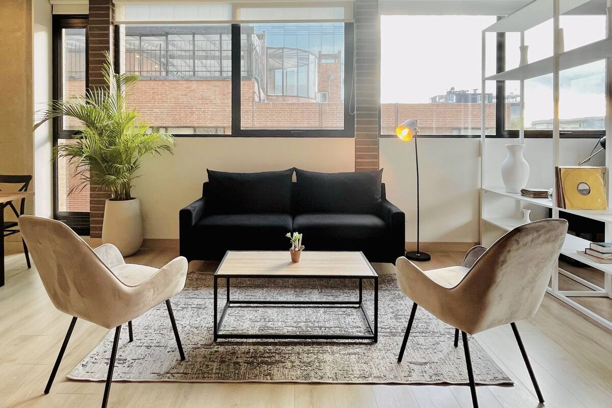 A modern living room with a black sofa, two armchairs, a coffee table, a potted plant, and a bookshelf.