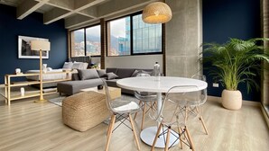 A modern living room with a round dining table, clear chairs, a sofa, a large potted plant, and a city view through large windows. - Canvas by Wynwood House (Bogotá)