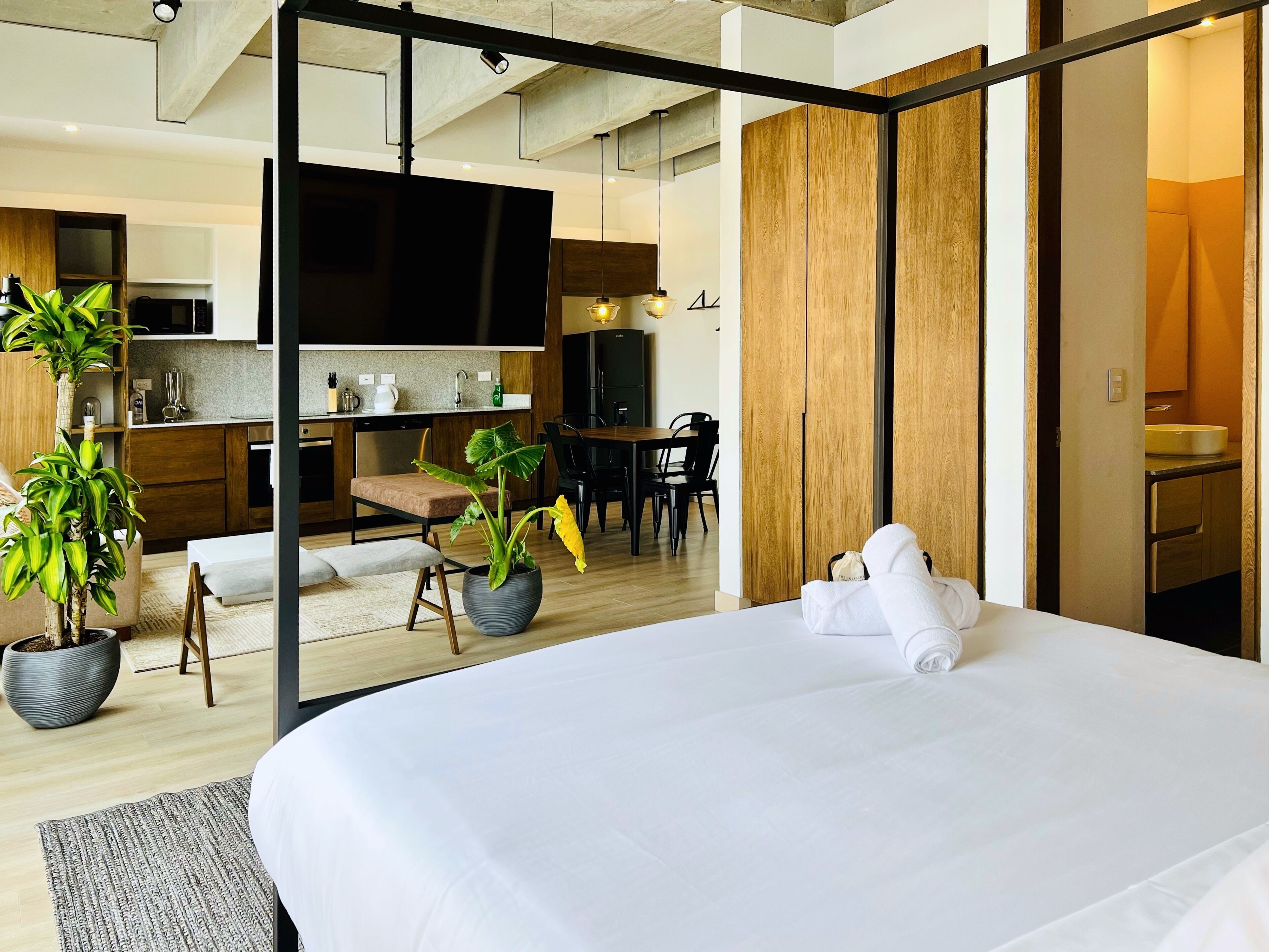 Habitación de hotel moderna con una cama grande, un área de comedor y una cocina visible a través de una puerta abierta.