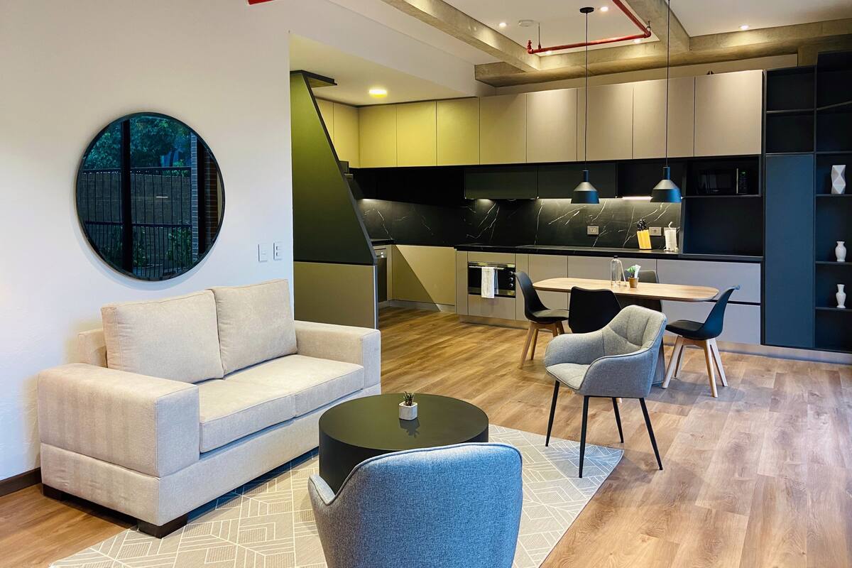 A modern living room with a sofa, chairs, a coffee table, and a circular mirror. There is a kitchen area with a dining table and chairs.