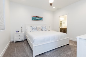Une chambre à coucher avec un lit blanc, une table de chevet avec une horloge, une horloge murale et une porte donnant sur une salle de bain.