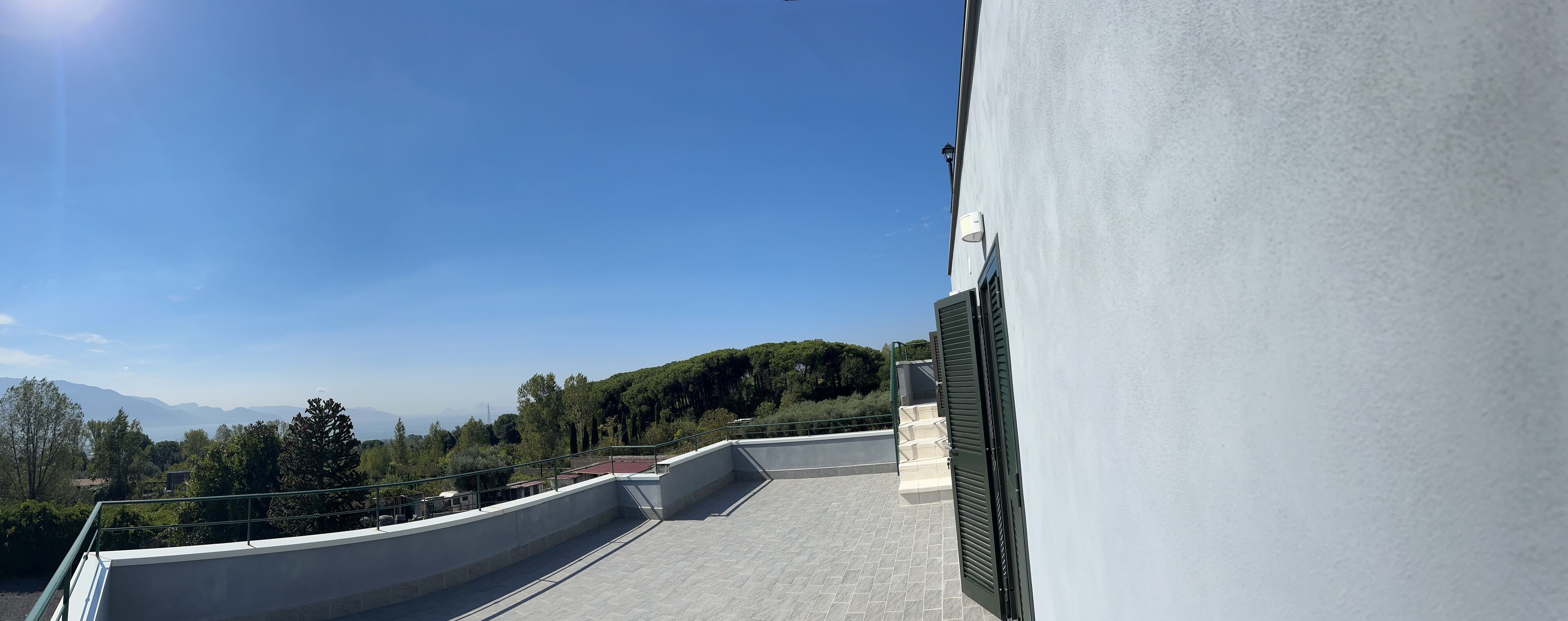 A rooftop terrace with a clear view of a mountainous landscape, a white building wall, and a metal railing.