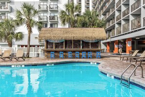 A hotel pool area with a thatched-roof bar, lounge chairs, and a multi-story building in the background. - Family Oceanfront Condo Located in North end of Myrtle Beach! (Myrtle Beach)