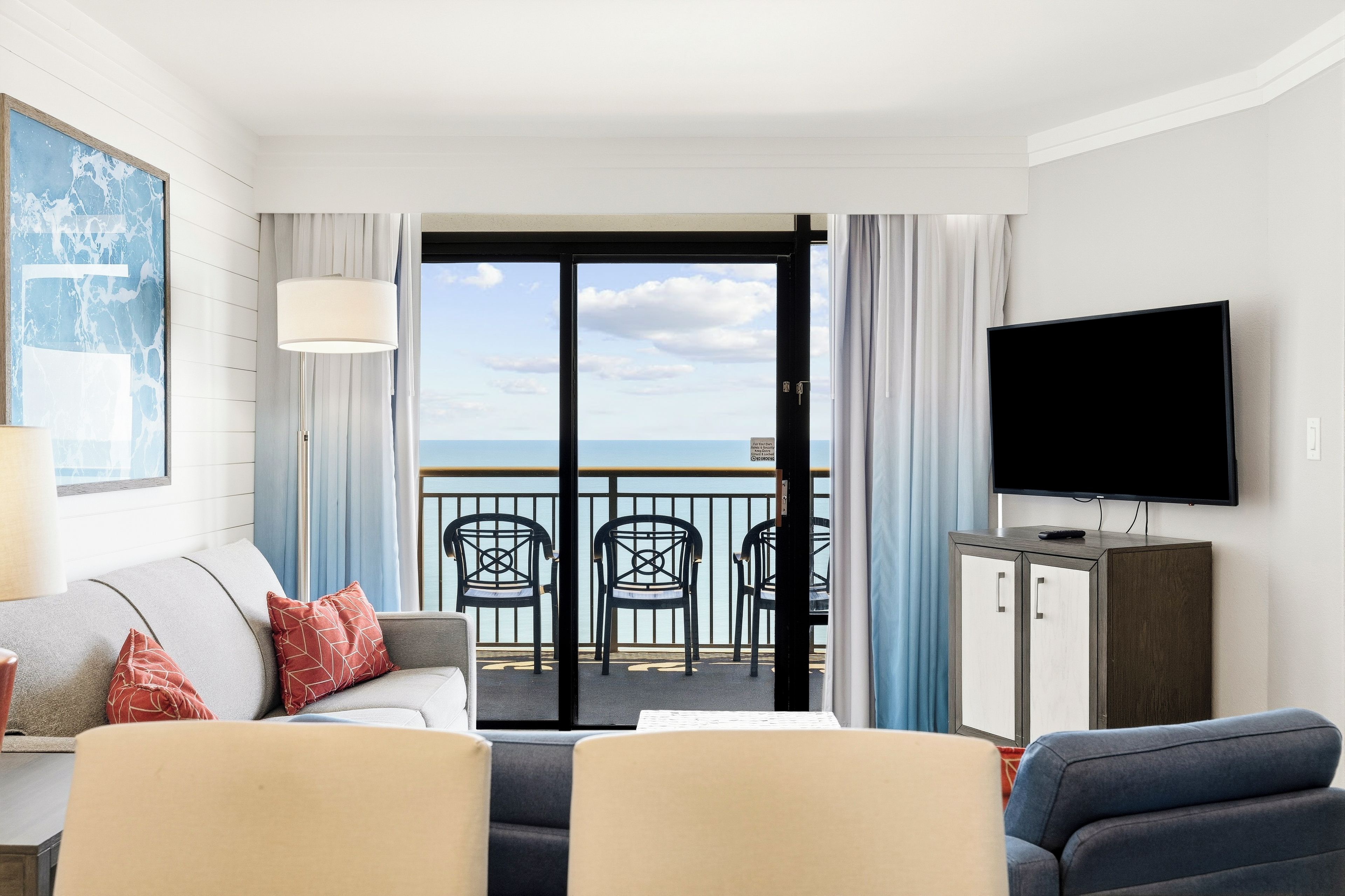 A hotel room with a balcony, a sofa, two armchairs, a television, and a cabinet.