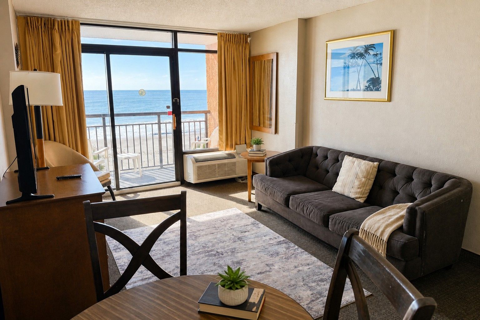 A room with a sofa, a table with a plant, a TV, and a view of the ocean through large windows.