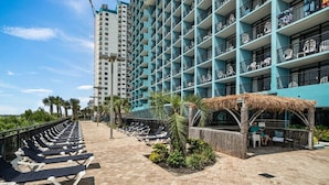 Un hotel con varios balcones, un área de piscina con tumbonas y una estructura con techo de paja.