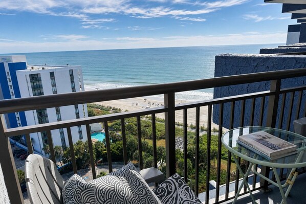 A balcony with a view of the ocean, a pool, and a building with a blue and white facade.