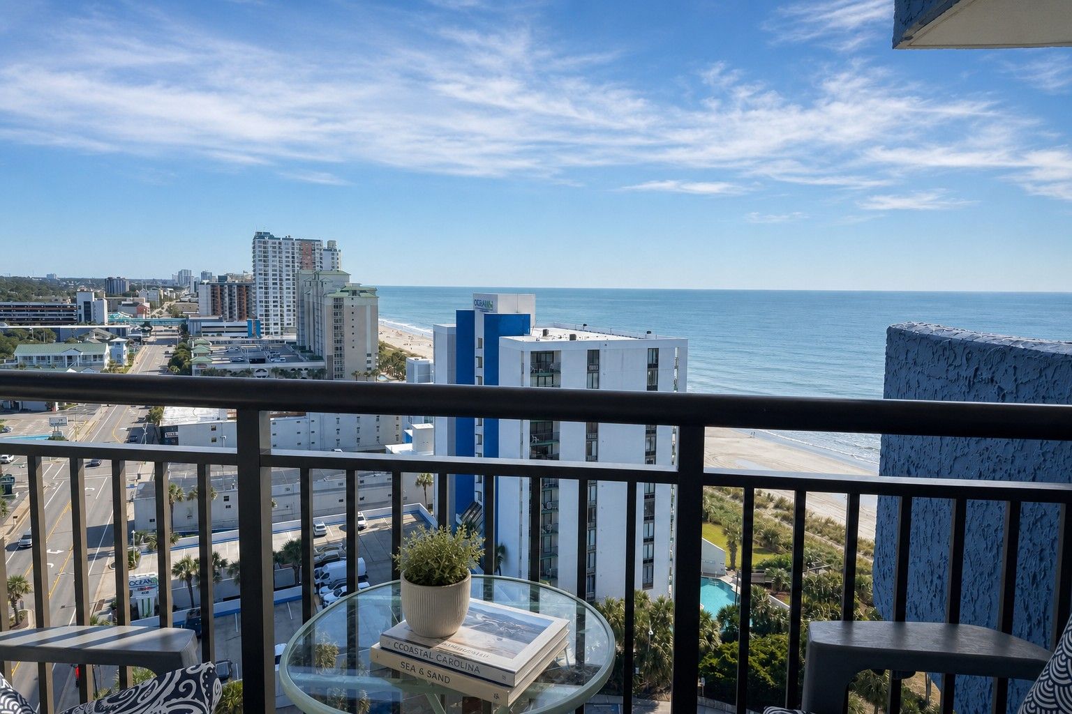 A balcony with a view of the ocean and cityscape, a potted plant, and a glass table with books.