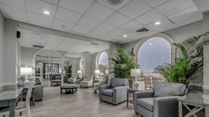 Eine geräumige Lobby mit grauen Sofas, einem Couchtisch und einem Essbereich mit Tisch und Stühlen. Große Fenster und Pflanzen prägen den Raum.