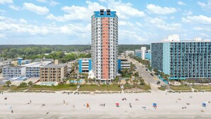 Ein Hochhaus-Hotel mit mehreren Etagen, einem Schwimmbad und einem Strandbereich mit Sonnenschirmen.