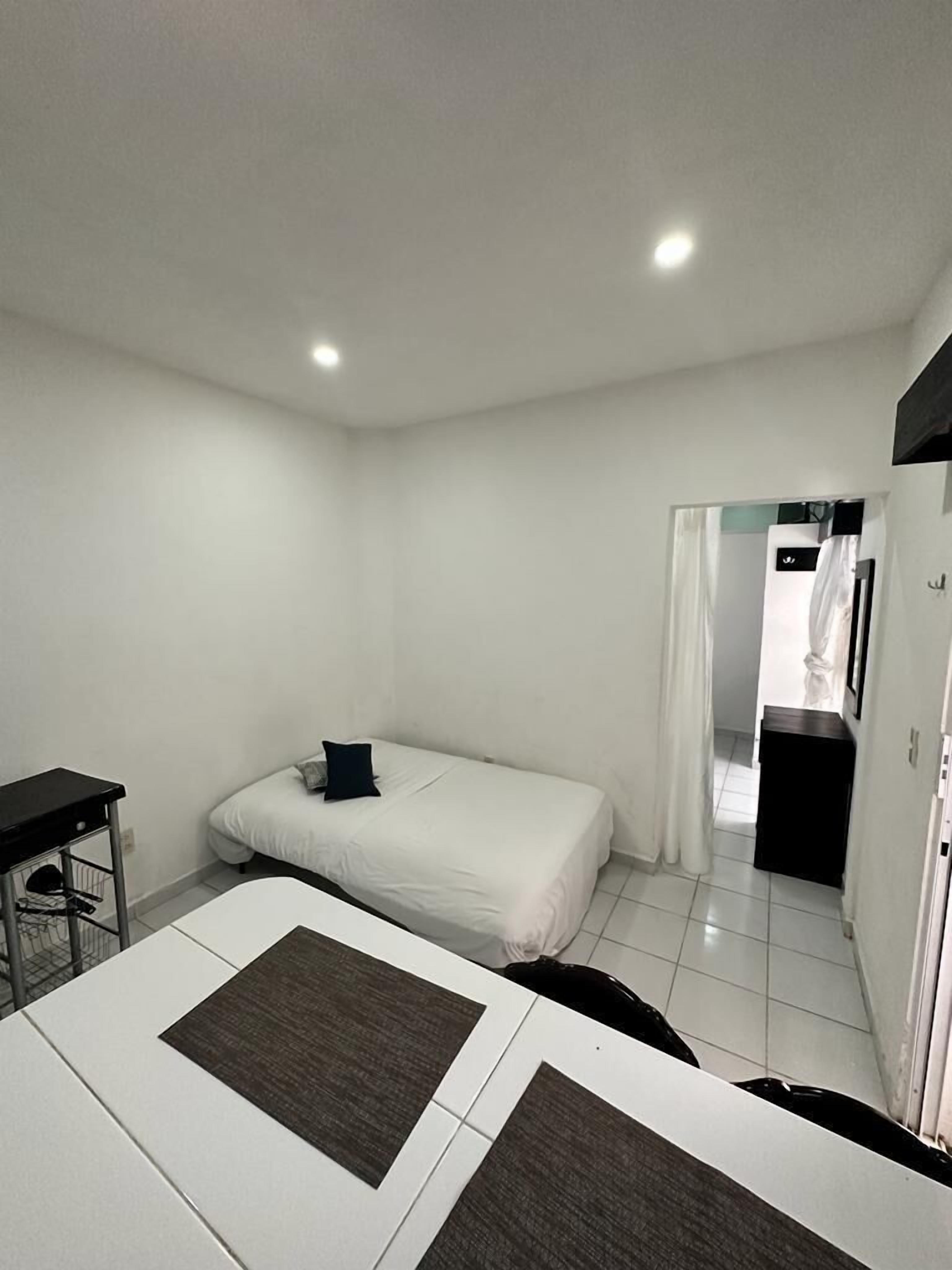 A small, clean room with a bed, a desk, and a chair. There is a door leading to another room.