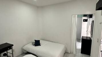 A small, clean room with a bed, a desk, and a chair. There is a door leading to another room.