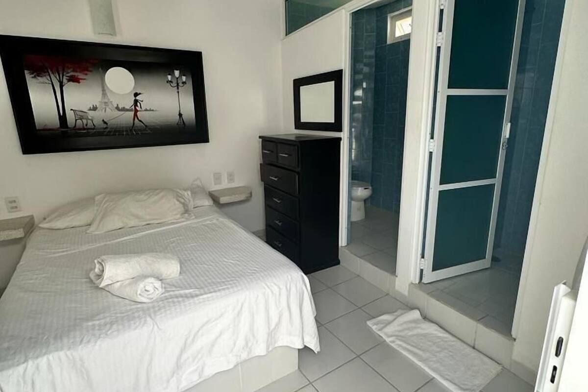 Un dormitorio moderno con una cama grande, una mesita de noche, un aparador, un baño con ducha y una puerta de cristal que da a un espacio exterior.