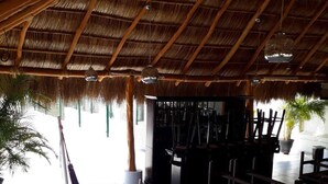 Un espacio al aire libre con techo de paja, una hamaca, asientos y plantas en macetas.