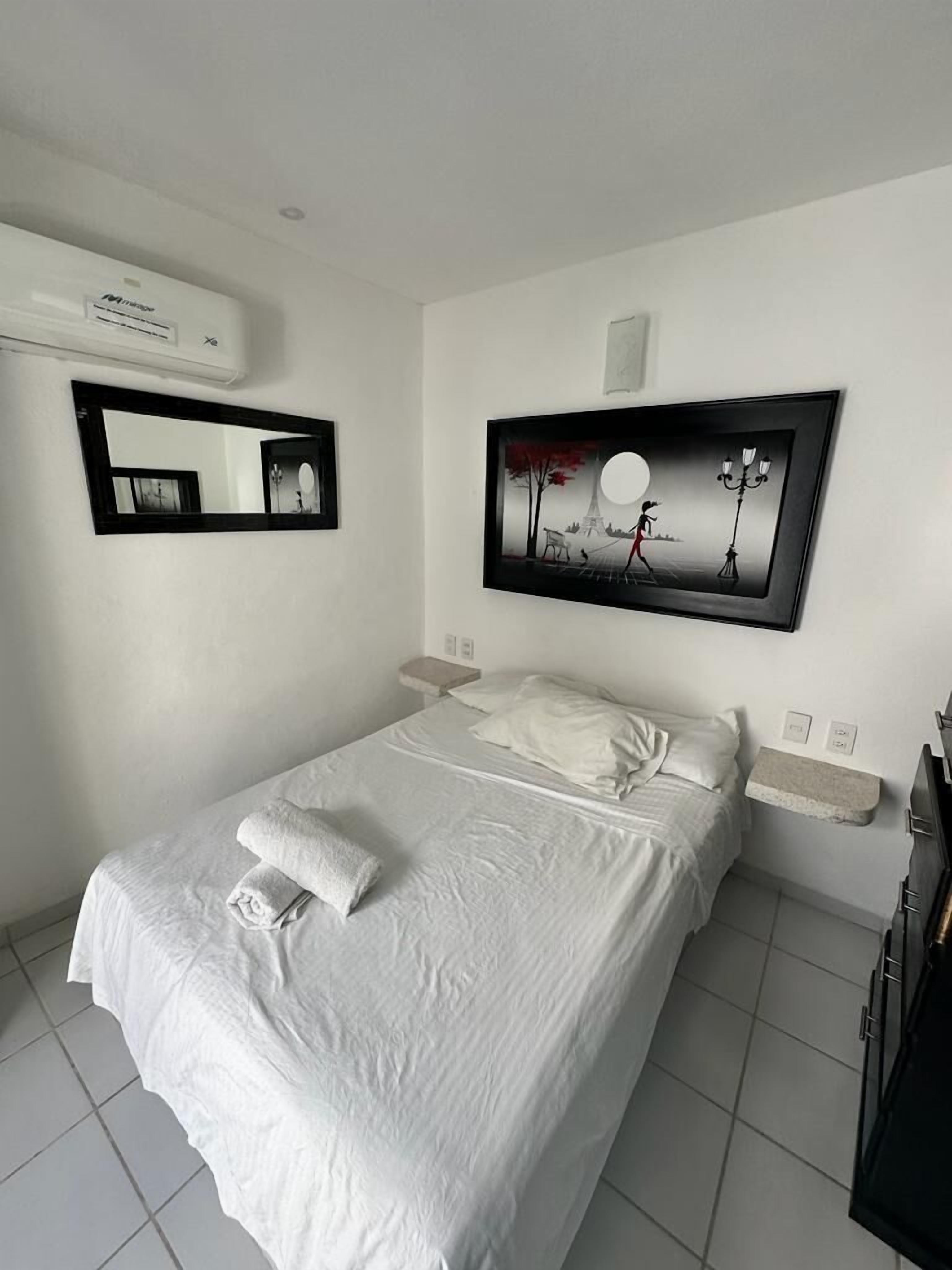 A neatly made bed with white linens and a folded towel, a wall-mounted air conditioner, a framed artwork depicting a night scene, and a small bedside table with a lamp.