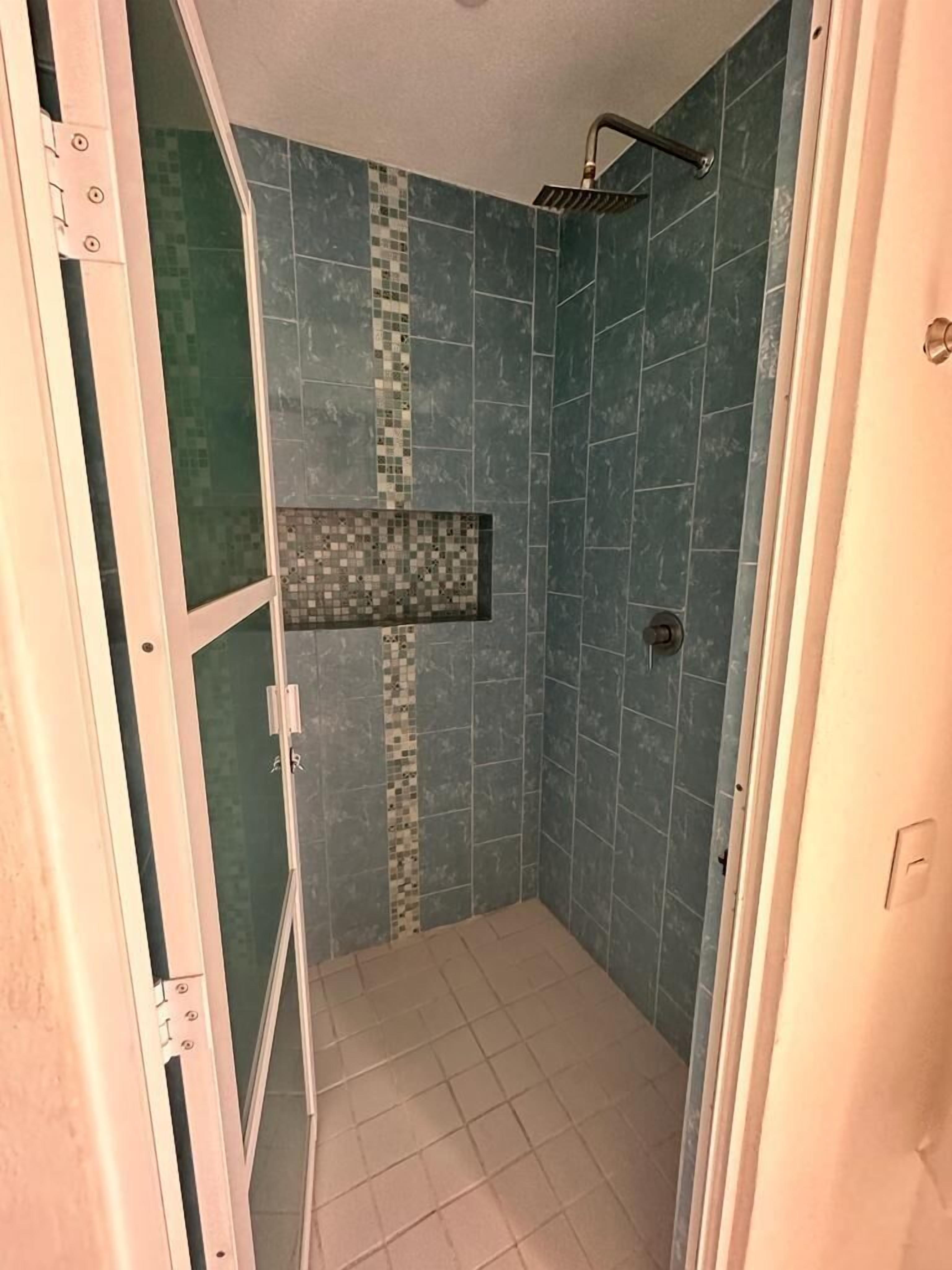 Un área de ducha pequeña y cerrada, con piso y paredes de azulejos, un cabezal de ducha y un estante pequeño con diseño de mosaico.