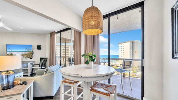 Una sala de estar moderna con una mesa redonda, taburetes de bar, un sofá y un amplio ventanal con vista al océano y edificios.