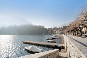 A lakeside view with boats docked, a stone wall, and buildings in the background.