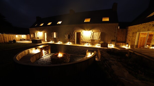 A stone building with a circular outdoor seating area and a fire pit, illuminated by warm lighting. - Gite Finistère for 7: Private Pool & Spa, Garden, 4 km from Beaches (PEUMERIT)