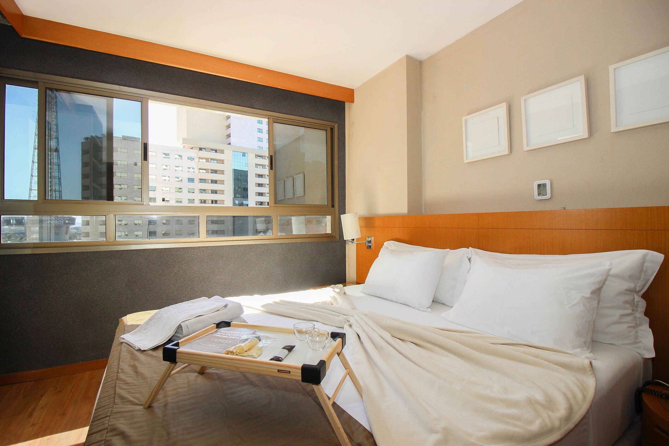 A hotel room with a large window, a bed with white linens, a bedside table, and a tray with items on it.