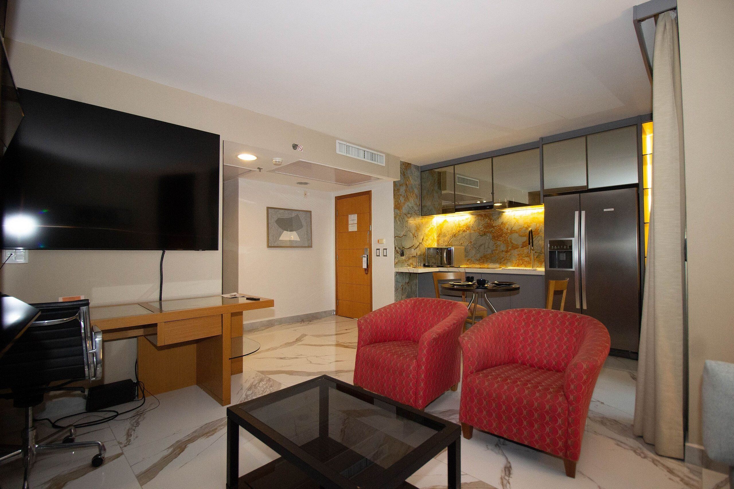Uma sala de estar moderna com televisão de tela plana, uma escrivaninha de madeira, uma mesa de centro de vidro e duas cadeiras vermelhas com padrão.