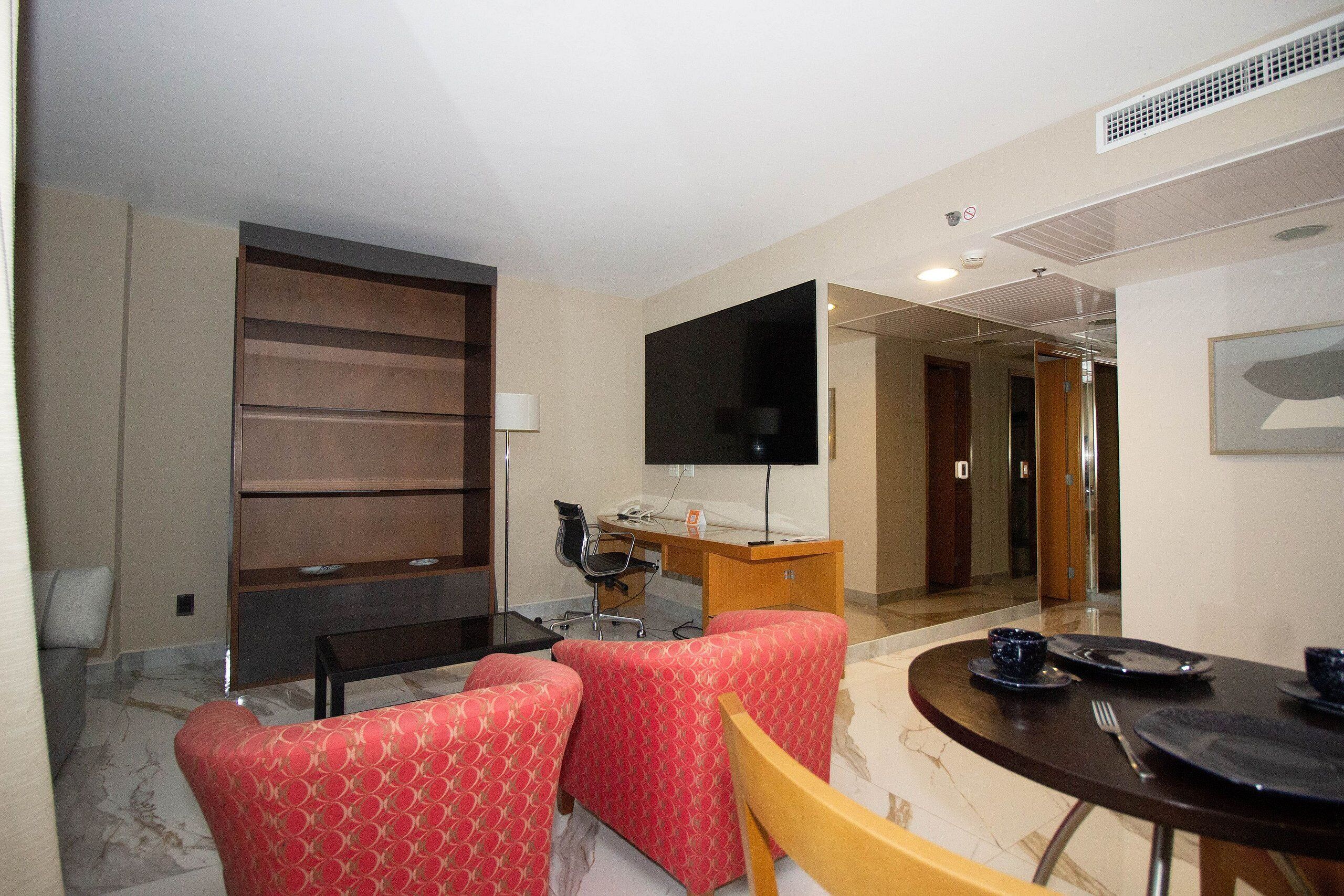Quarto de hotel com sofá, escrivaninha, televisão e mesa de jantar posta para dois.
