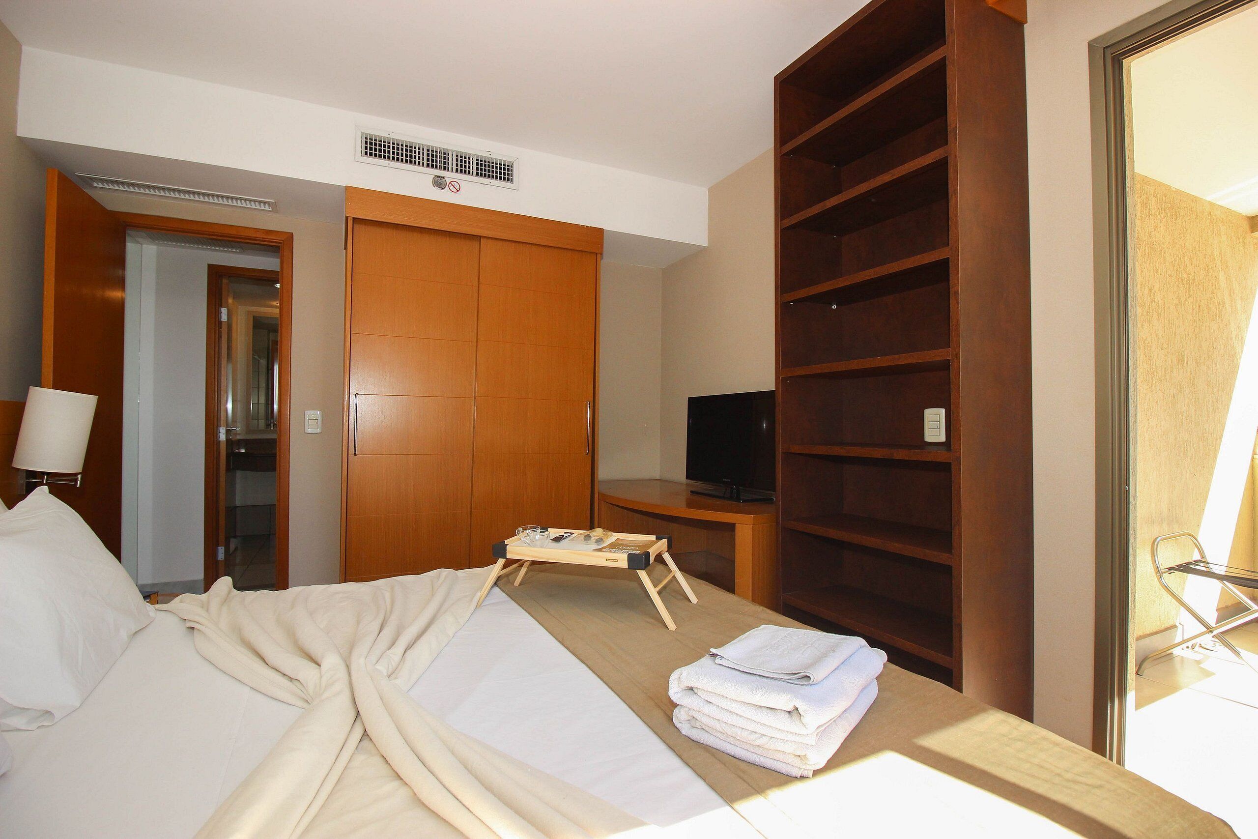 A hotel room with a bed, a TV, a wardrobe, and a shelf with folded towels.