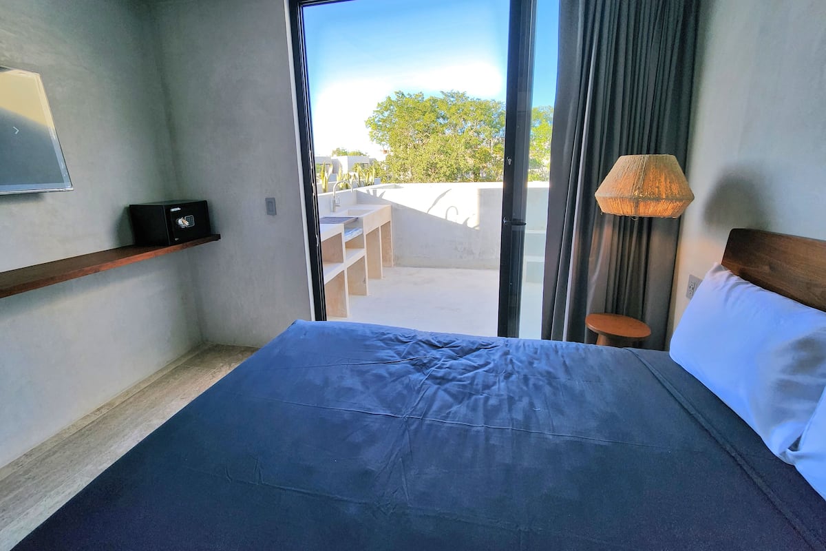 A hotel room with a bed, a TV, a small table, a lamp, and a view of the outdoors.