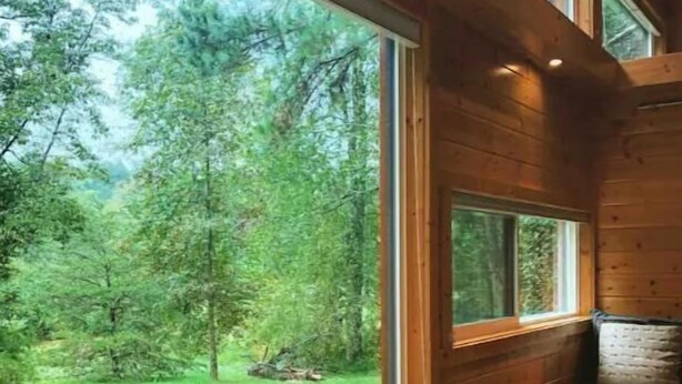 Une cabane en bois comprenant une table et des chaises, un canapé et de grandes fenêtres donnant sur une zone boisée.
