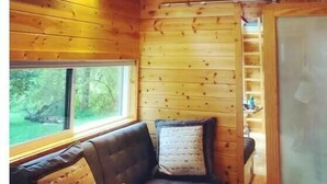 L’intérieur d’une cabane avec des murs en bois, un espace en mezzanine, une fenêtre donnant sur la forêt et un ventilateur de plafond.