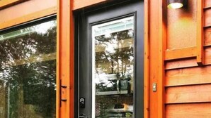 Une façade en bois, avec une porte et des fenêtres en verre, qui reflètent les arbres et l’intérieur d’une cuisine.
