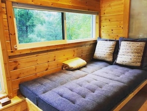 A cozy cabin interior with a wooden wall, a window with a view of trees, a cushioned bench with pillows, and a small wooden table. - Your Perfect Blue Ridge Mountain Hideaway near 151 (Afton)