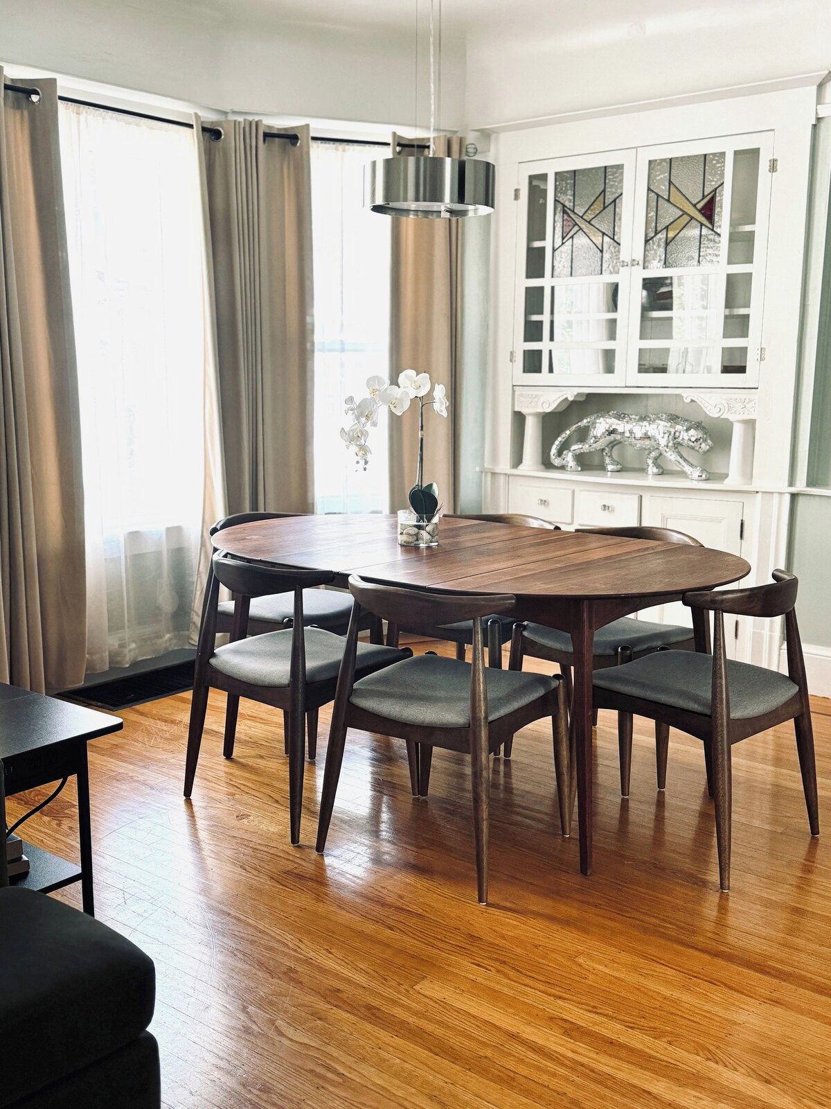 A dining area with a round wooden table and six chairs, a hanging light fixture, and a decorative glass cabinet with a metal sculpture.