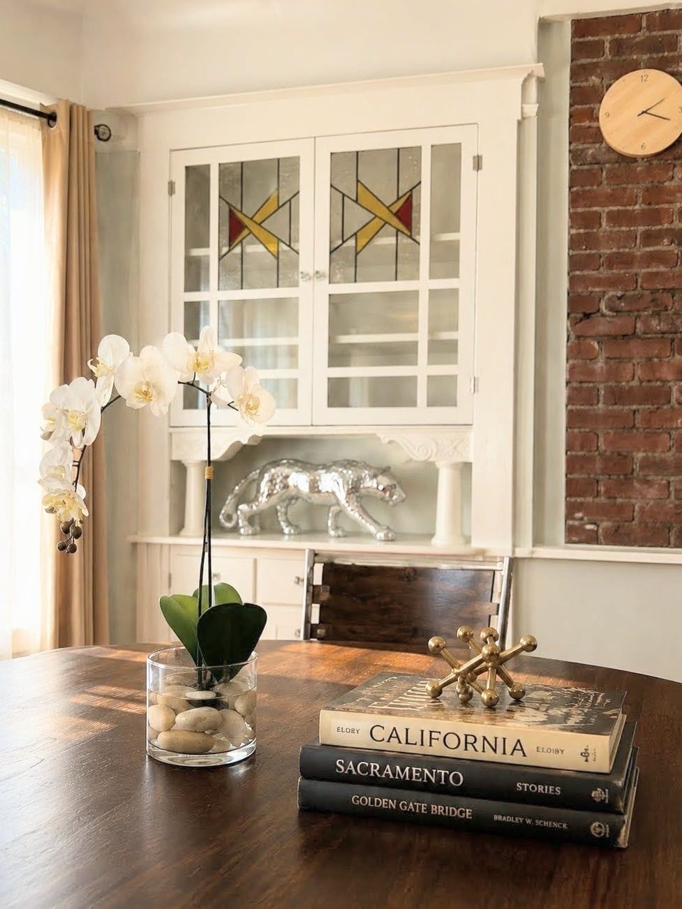 Una mesa de comedor con un jarrón de vidrio con orquídeas blancas, una pila de libros y una pieza decorativa de metal.