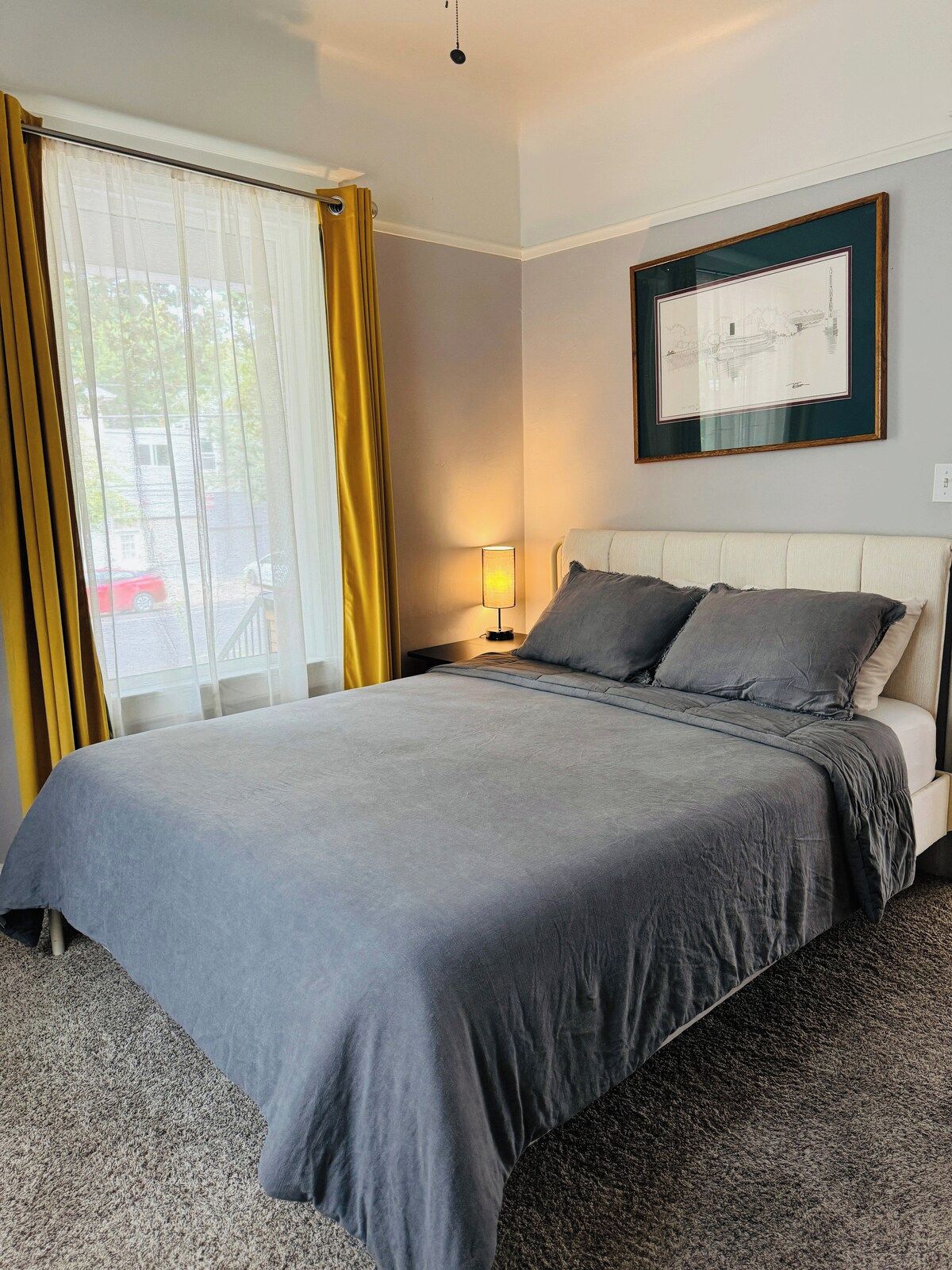 Una camera d'hotel con un letto, un comodino, una lampada, un quadro appeso al muro e una finestra con tende.