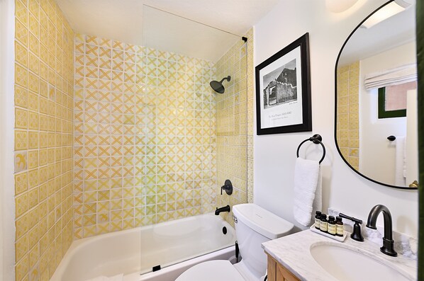Un baño con baldosas amarillas estampadas, una bañera, un inodoro, un lavabo y un cuadro en la pared.