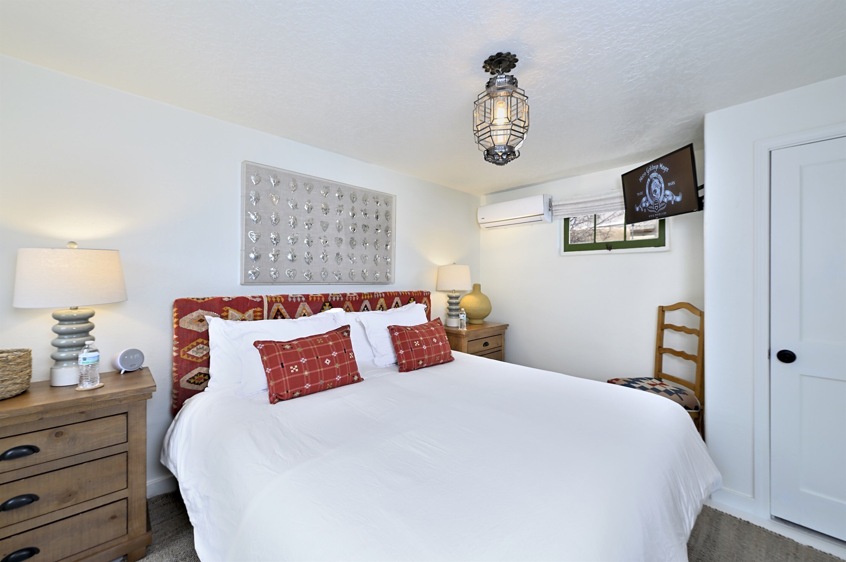 A neatly made bed with white bedding and red pillows, a wooden nightstand with a lamp, a wall clock, and a decorative wall panel.