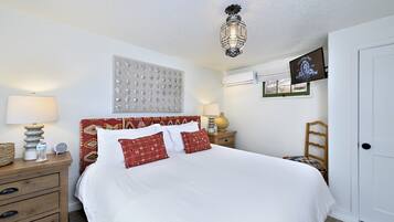 A neatly made bed with white bedding and red pillows, a wooden nightstand with a lamp, a wall clock, and a decorative wall panel.