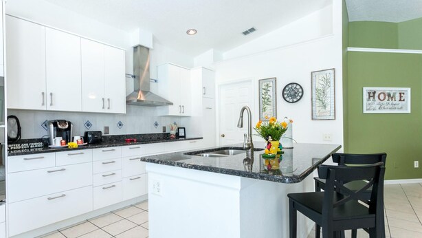 A modern kitchen with white cabinets, a granite countertop, a stainless steel sink, and a black chair. - Free Pool Heat & Spacious Pool Deck | Themed Bedrooms | Near Disney (Kissimmee)