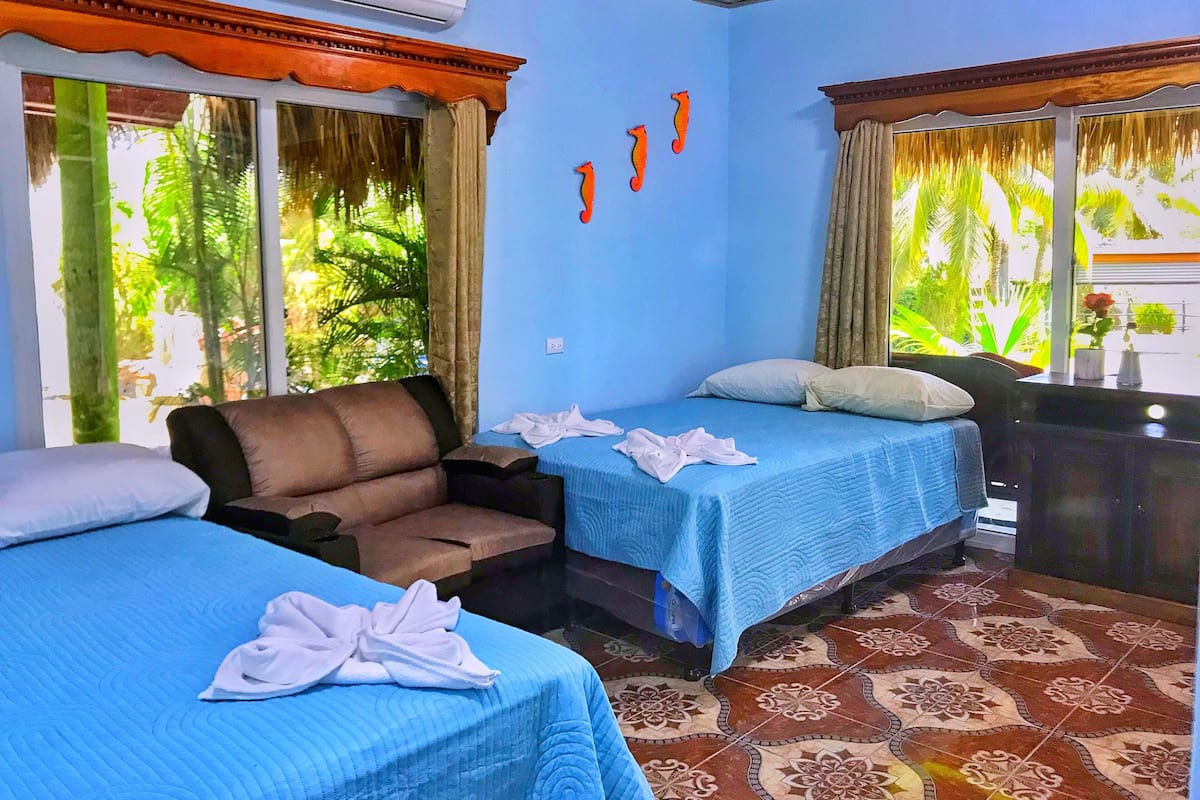 A hotel room with two beds, a sofa, and a window with a view of greenery.